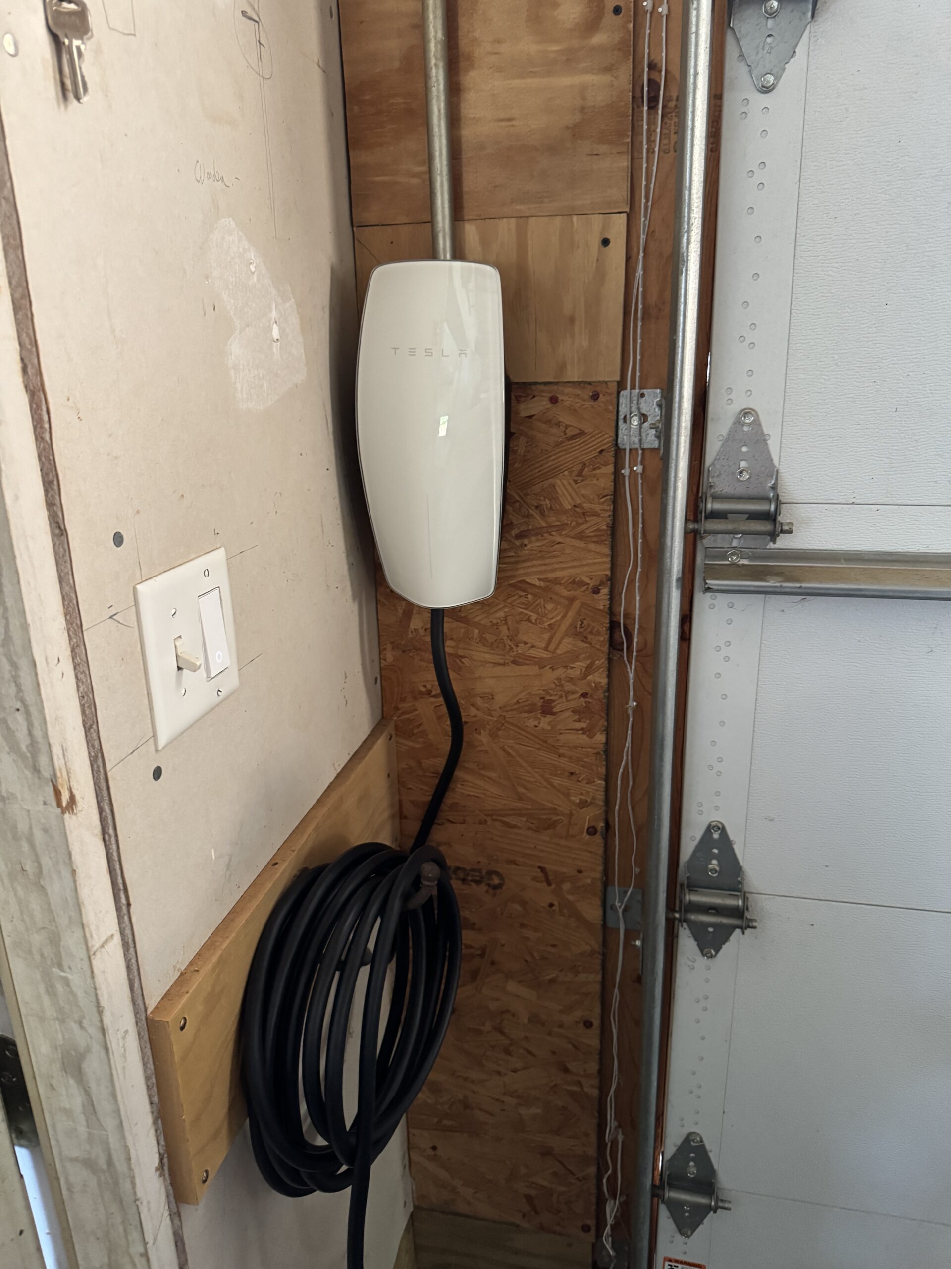 A Tesla wall connector, part of modern EV charging stations, is mounted on a wooden panel in a garage, with its charging cable neatly coiled below.