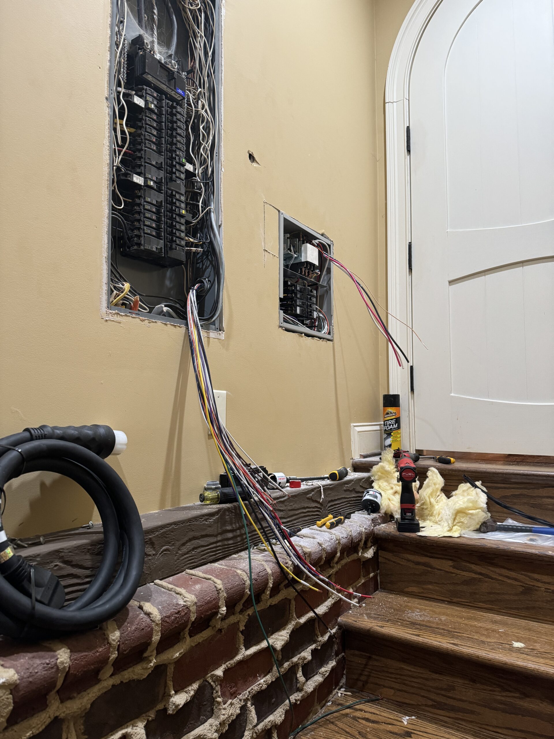 Two open electrical panels with exposed wires beside a staircase; tools and materials are scattered on the wooden steps and brick wall.