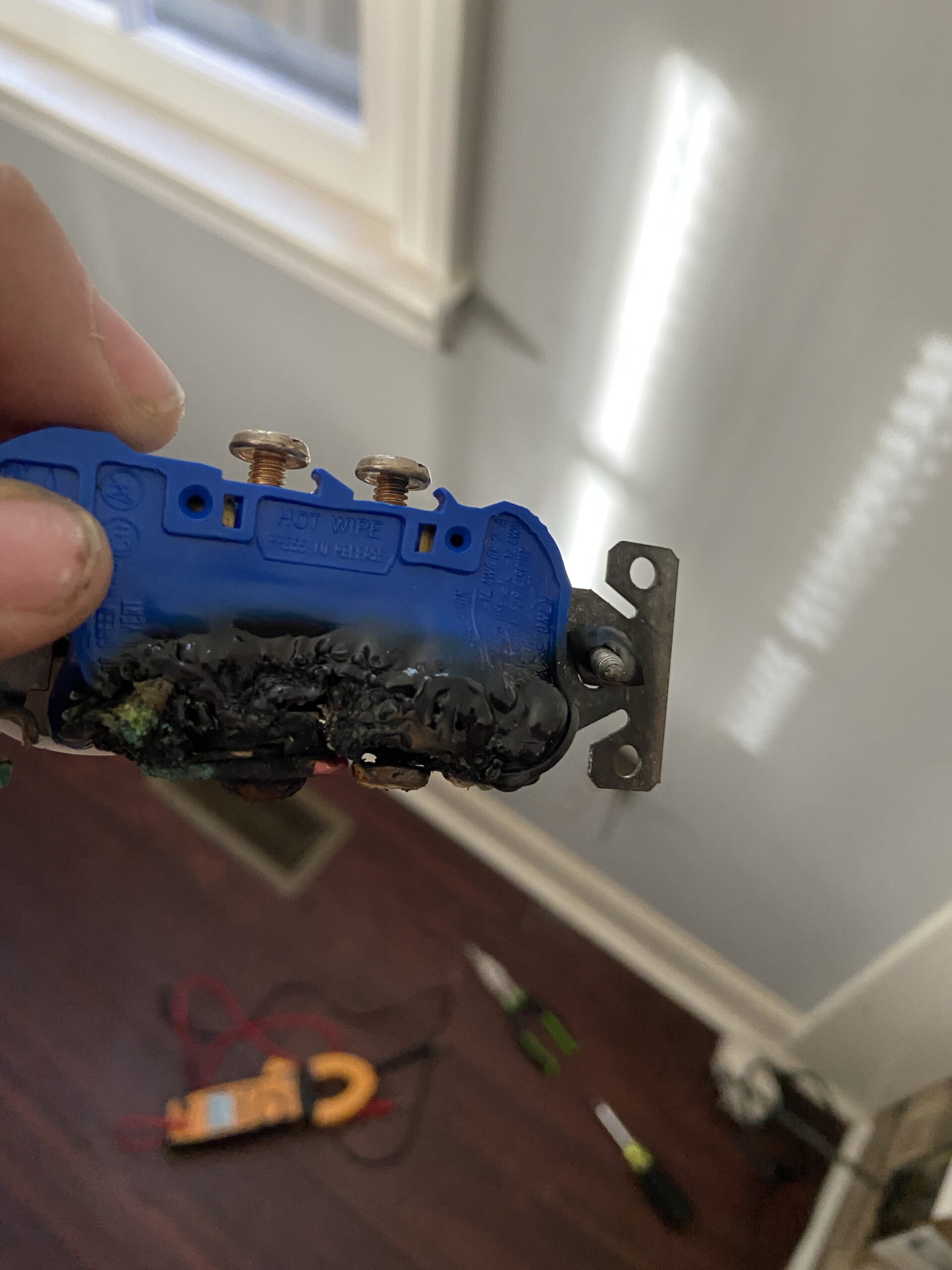 A hand holds a damaged electrical outlet with melted and burnt plastic; tools are visible on the floor in the background.