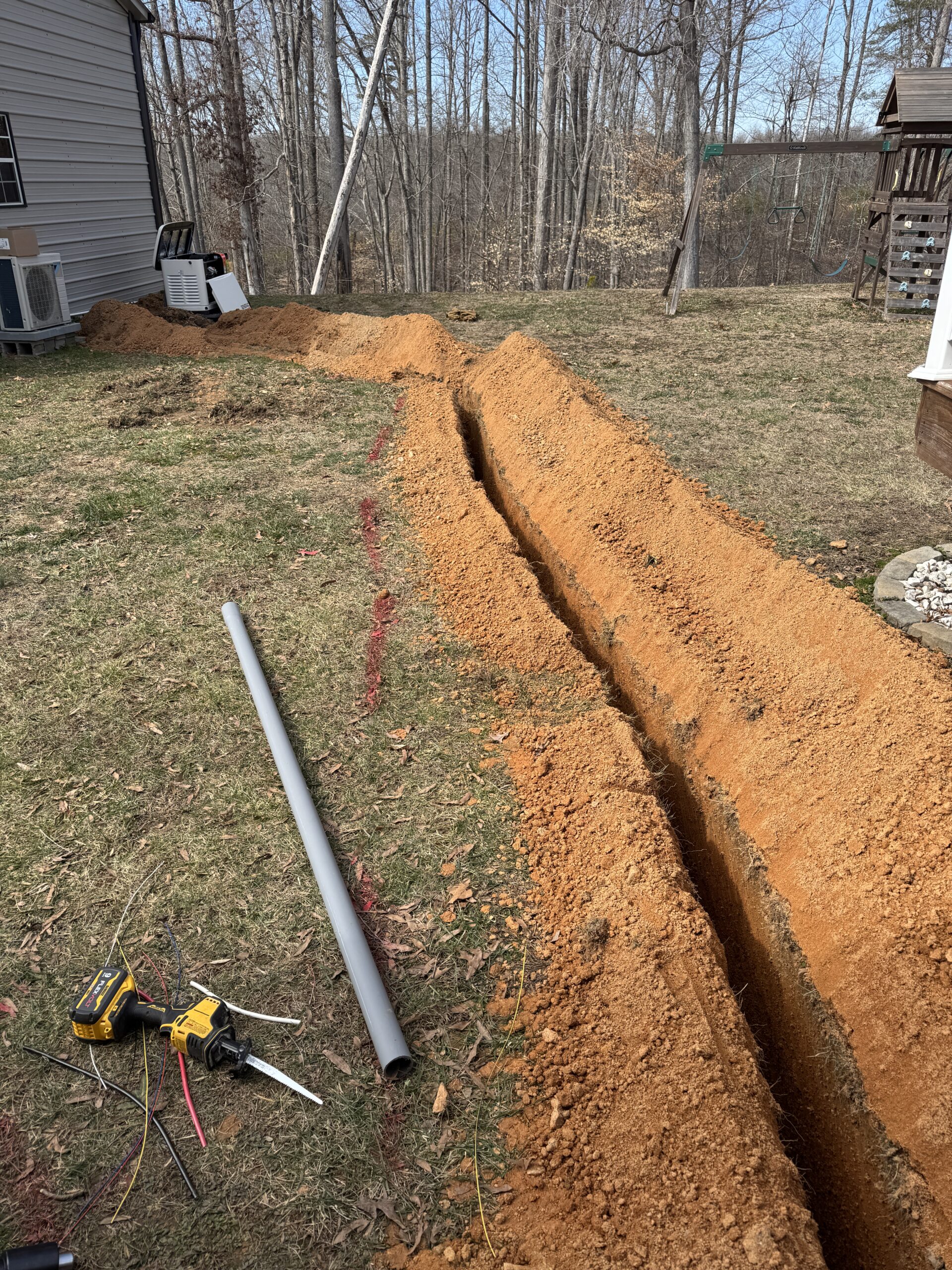 A narrow trench dug through a yard with exposed soil, red guide lines, a gray pipe, power drill, wires, and tools scattered nearby. Trees and buildings are in the background.