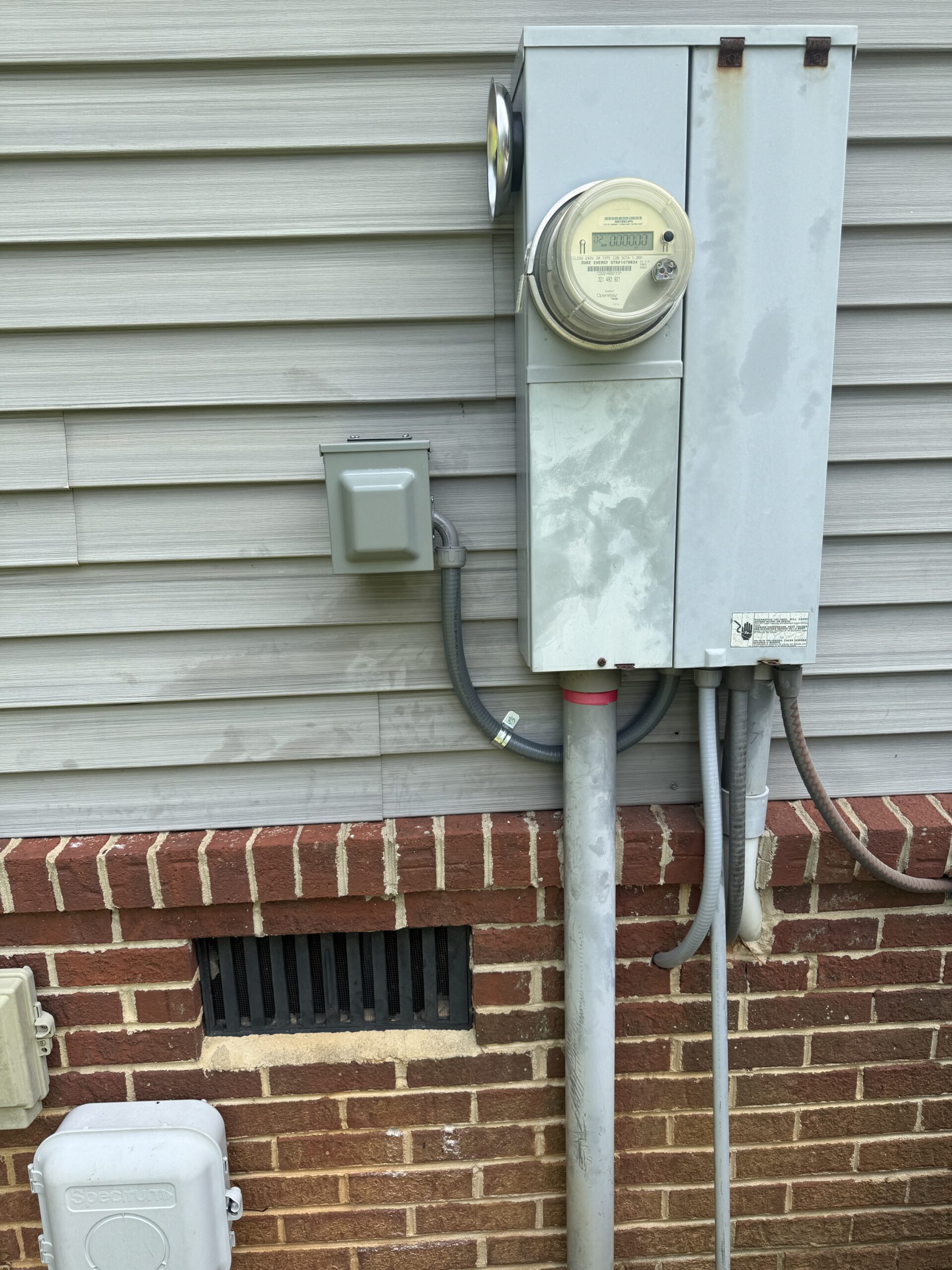 An electric meter and breaker box mounted on the exterior wall of a house with vinyl siding and a brick foundation.
