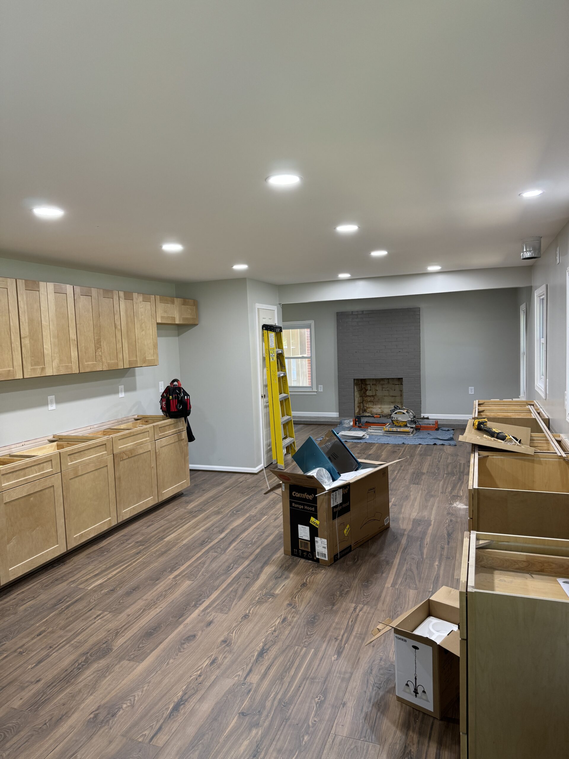 A kitchen under construction with cabinets installed, a ladder near the fireplace, scattered boxes, and tools on a wood floor.