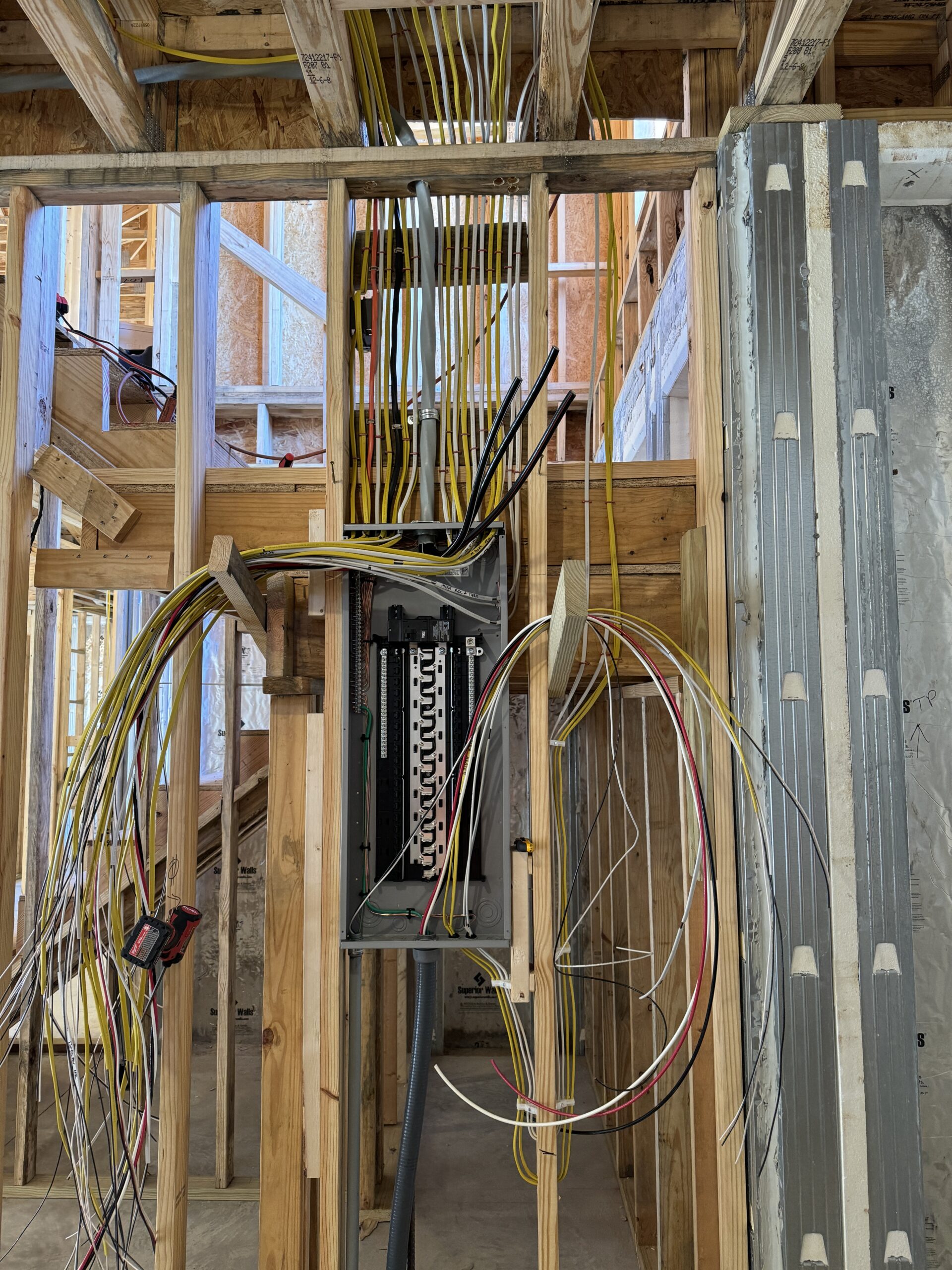 Exposed electrical wiring and open circuit breaker panel in a wooden stud wall of a building under construction.