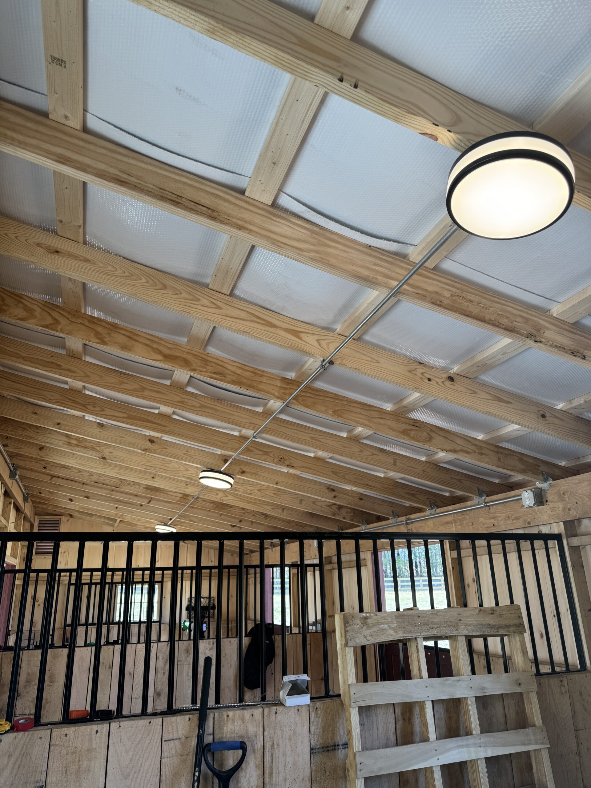 Interior of a wooden stable with exposed beams, lit ceiling lights, black metal stall bars, and stable equipment visible.