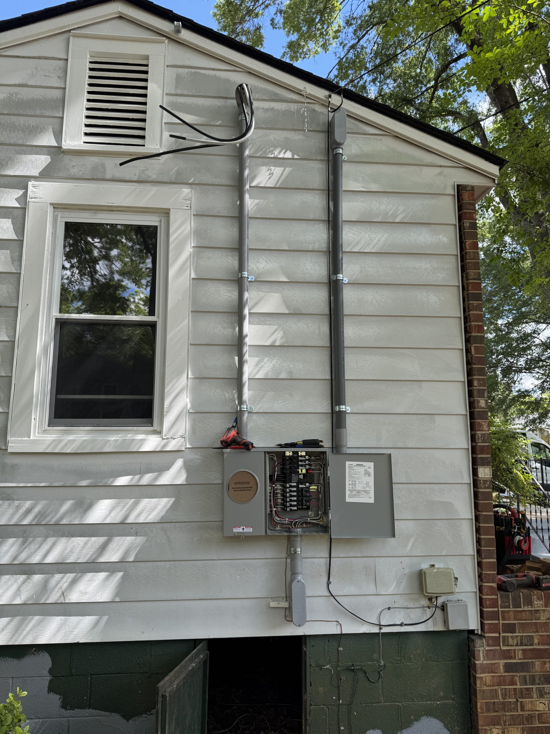 Two metal electrical conduits run vertically up the side of a white house, connecting to an open electrical panel and various utility boxes, with cables exposed at the top.