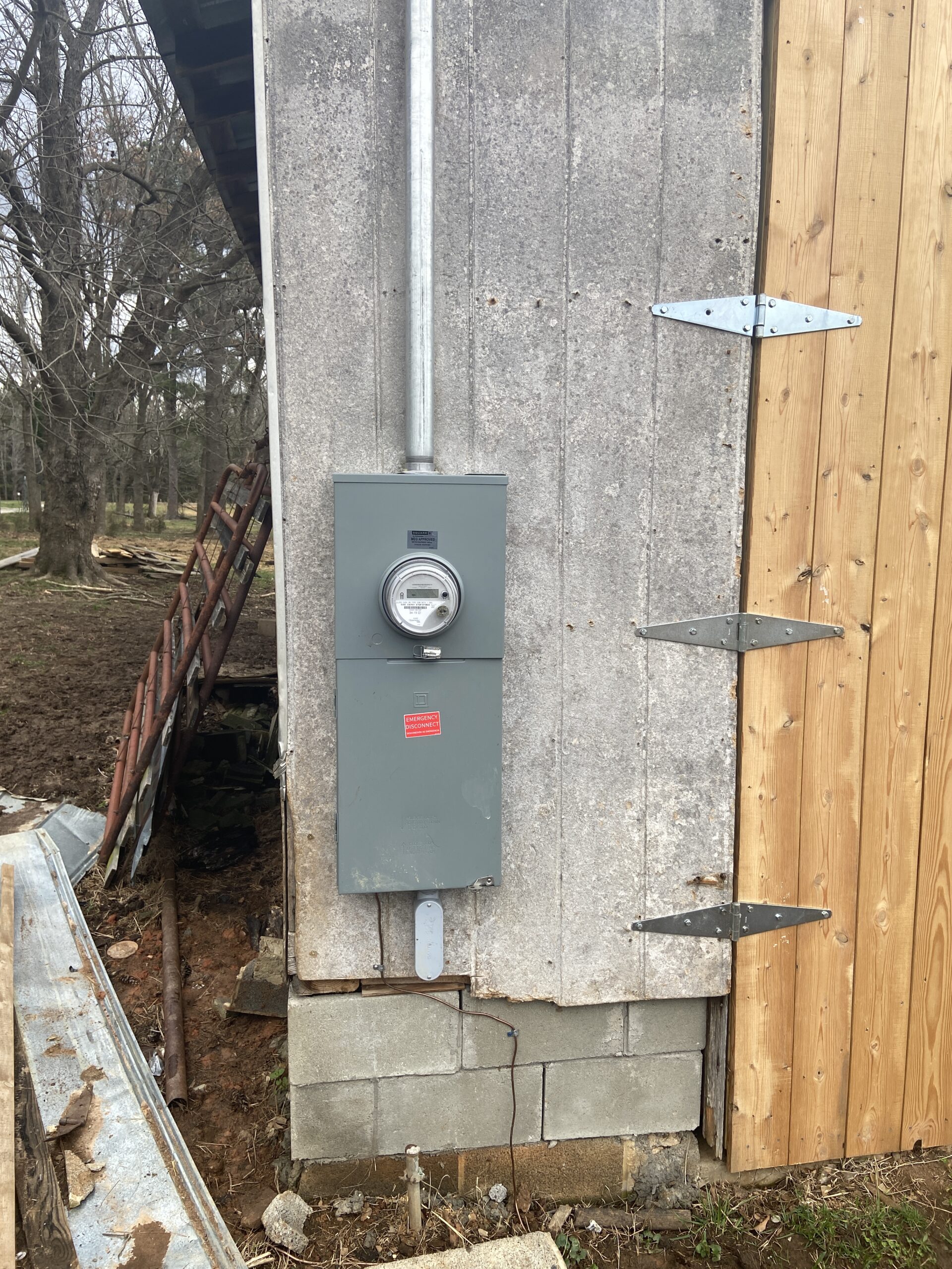 An electric meter box mounted on an exterior concrete wall corner, next to a new wooden structure with large metal brackets.