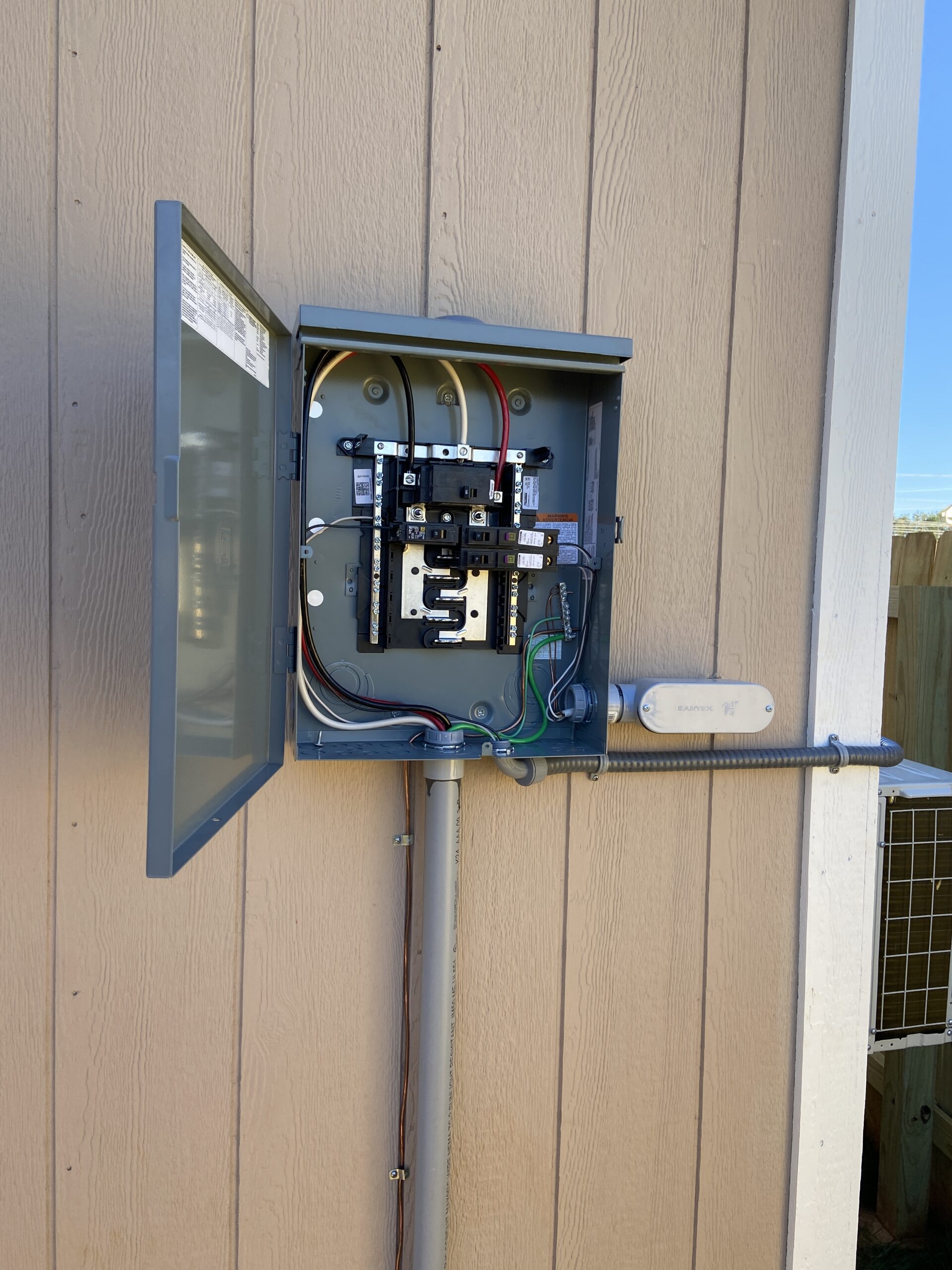 An open outdoor electrical breaker panel mounted on a wooden wall, showing exposed wiring and circuit breakers inside the metal box.